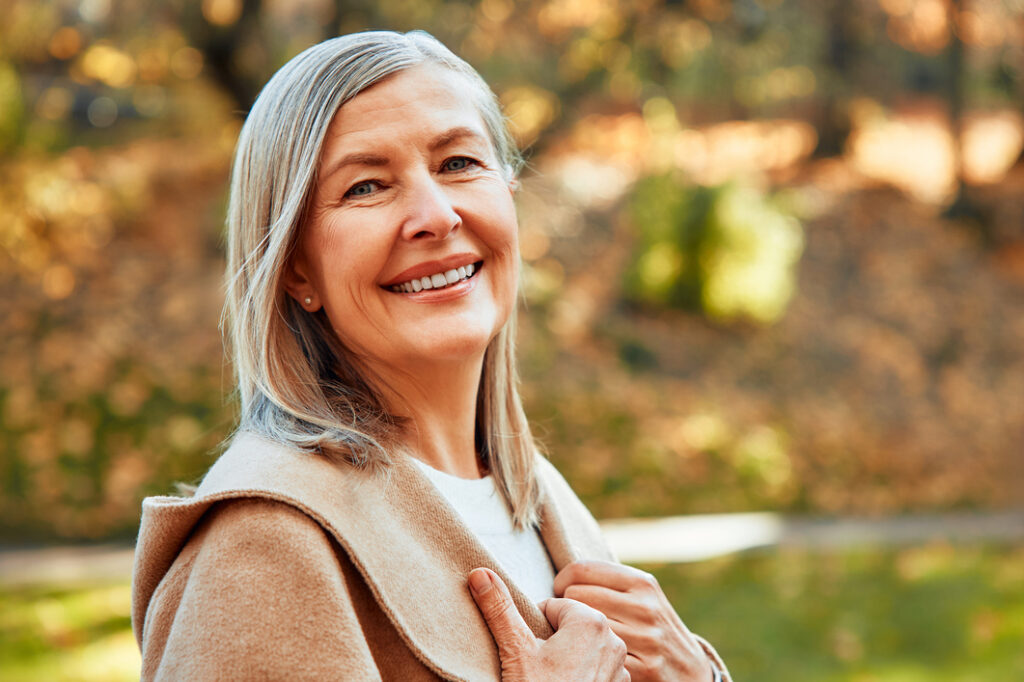 Senior woman smiling outdoors at Vista Prairie at Fieldcrest senior living community in Sheldon, Iowa, enjoying autumn scenery and pleasant weather.