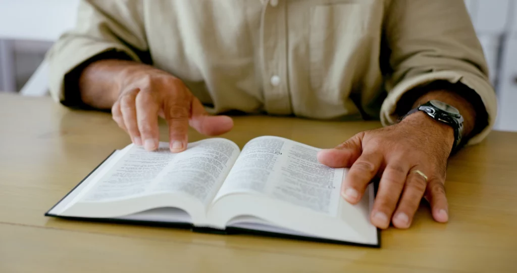 A person reading a Bible, close-up of hands, senior man at a wooden table, reading scripture, peaceful activity in a comfortable environment.