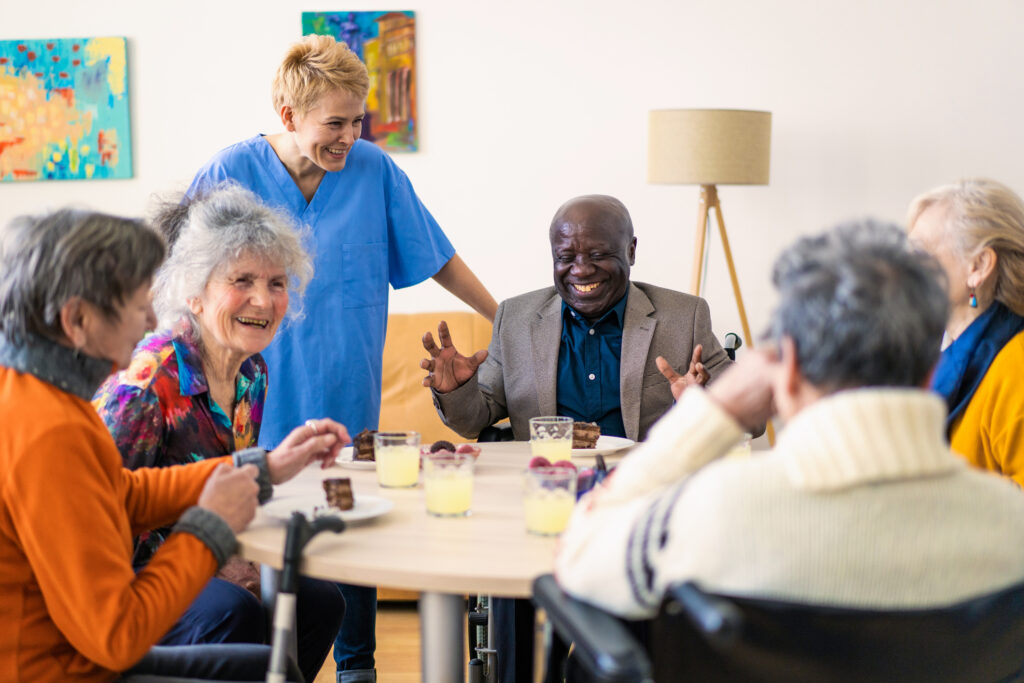 Friendly senior residents enjoying a social gathering at Vista Prairie at Windmill Ponds senior living community in Alexandria, MN with caring staff member smiling in the background.