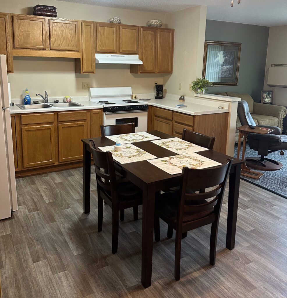 Modern senior living apartment kitchen at Vista Prairie Windmill Ponds, featuring a cozy dining area with a dark wood table and chairs, and a well-equipped kitchen with wooden cabinets and white countertops.