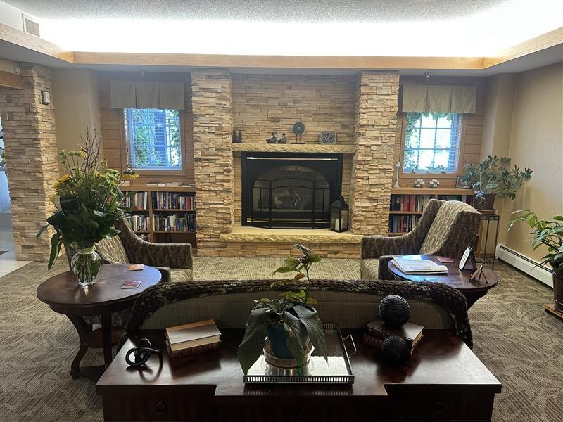Cozy senior living living room at Vista Prairie at Windmill Ponds in Alexandria, MN, featuring a stone fireplace, comfortable seating, and large windows with natural light for a welcoming atmosphere.