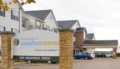 Comprehensive view of Vista Prairie at Goldfinch Estates senior living community's entrance, featuring a welcoming sign and multi-story residential buildings in Fairmont, Minnesota.
