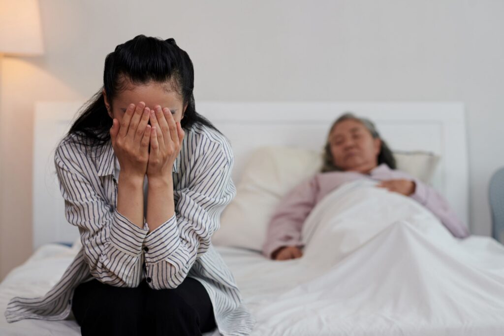 picture of younger woman sitting at end of bed with her head in her hands while older woman sleeps in bed