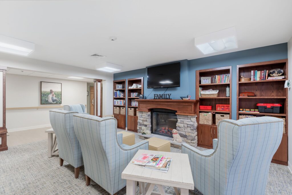 Cozy senior living lounge at Vista Prairie at Ridgeway in New Ulm, Minnesota, featuring a stone fireplace, comfortable armchairs, bookshelves, and a welcoming family atmosphere.