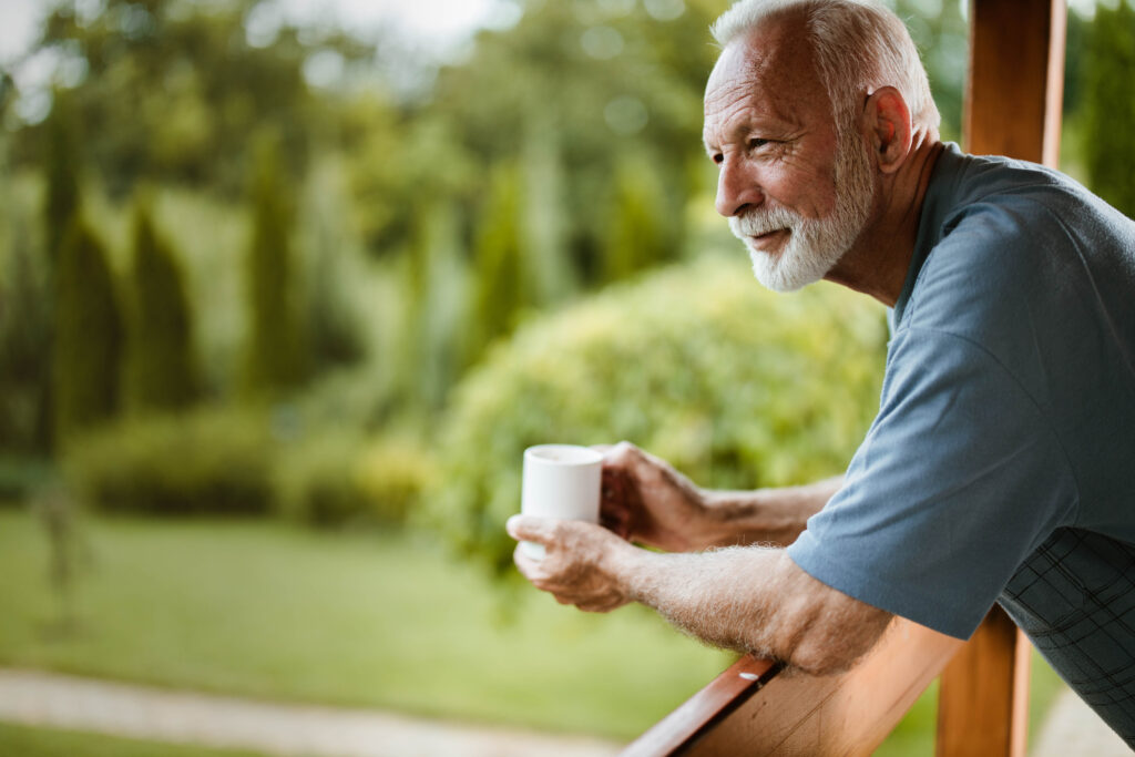 Peaceful senior man enjoying morning coffee on porch at Vista Prairie at Ridgeway senior living community in New Ulm, Minnesota, surrounded by lush greenery.