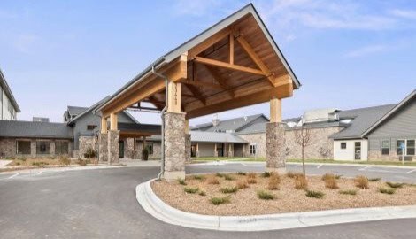 Modern senior living community entrance with a wooden and stone canopy, landscaped surroundings, and accessible parking in New Ulm, Minnesota.