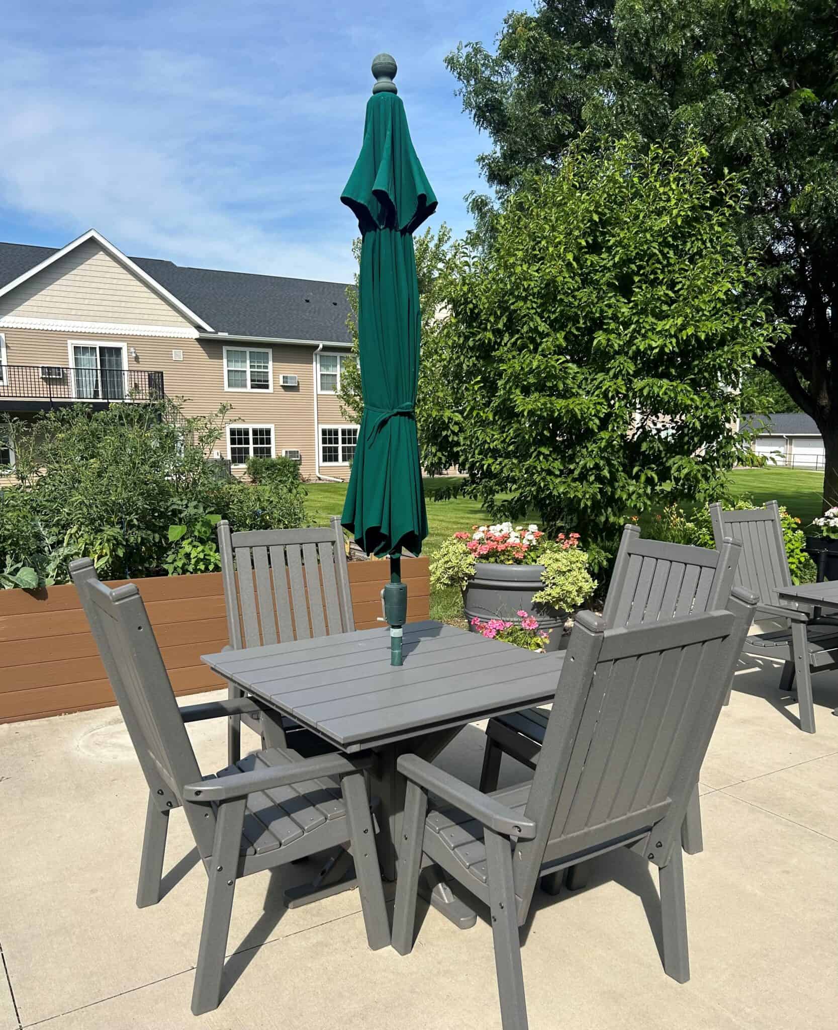 Outdoor senior living patio with gray tables, chairs, a closed green umbrella, lush trees, colorful flowers, and apartment buildings in the background at Vista Prairie North Mankato.