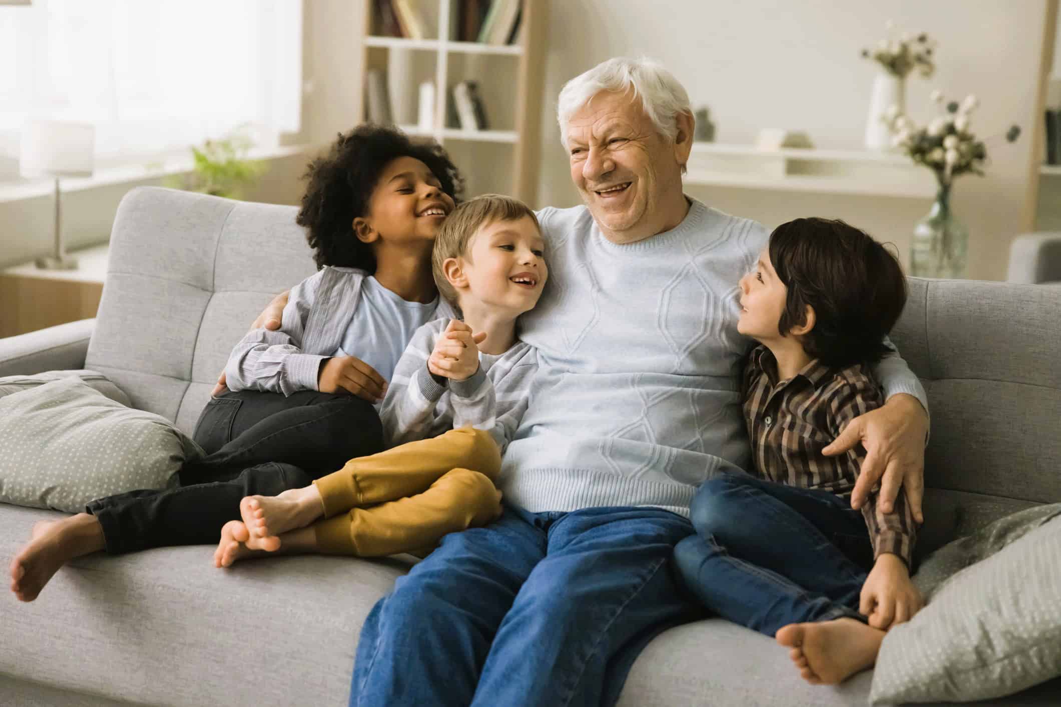 Happy elderly man sitting on a sofa surrounded by smiling children, enjoying a joyful moment together in a cozy, well-lit living room.