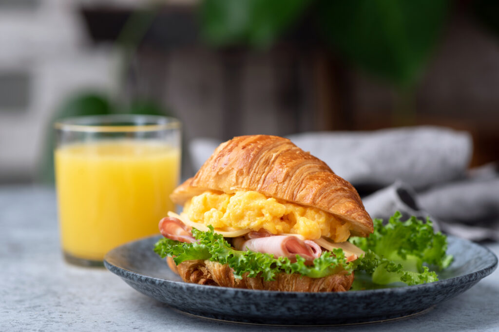 Fluffy scrambled eggs with ham on a buttery croissant served with fresh green lettuce, accompanied by a glass of orange juice, perfect for breakfast at Vista Prairie at Garnette Gardens.
