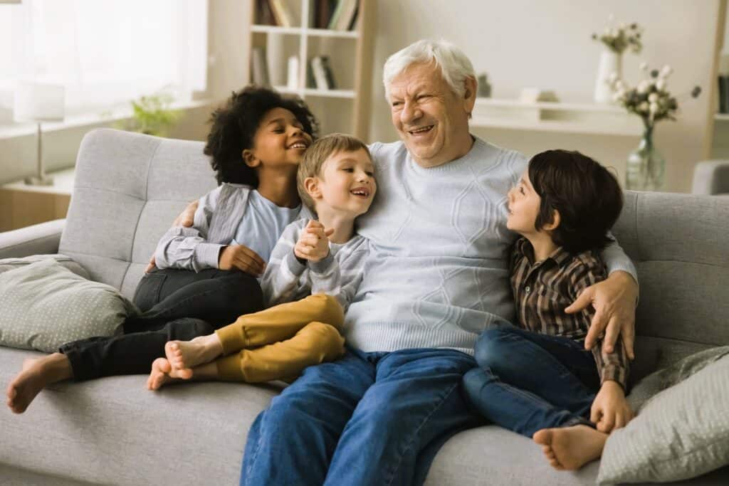 Senior man with grandchildren smiling and enjoying time together on a cozy gray sofa in a bright living room.
