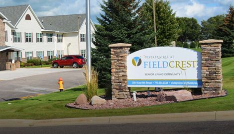 Bright, welcoming sign for Vista Prairie at Fieldcrest senior living in Redwood Falls, MN, featuring stone pillars and lush landscaping, offering a comfortable, friendly environment for seniors.