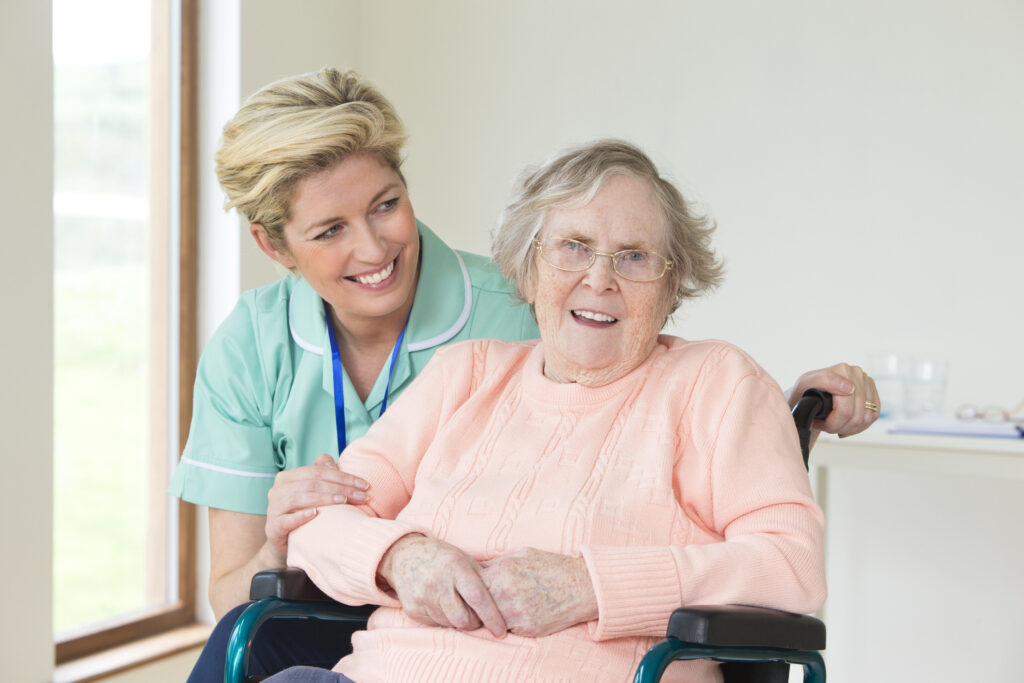 Elderly woman in wheelchair with caregiver smiling in senior living facility, showcasing compassionate elderly care and support at Vista Prairie at Eagle Pointe in St. Francis, MN.