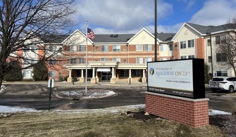 Senior living community building with a sign in front, featuring a brick base and a large digital display, under a partly cloudy sky.