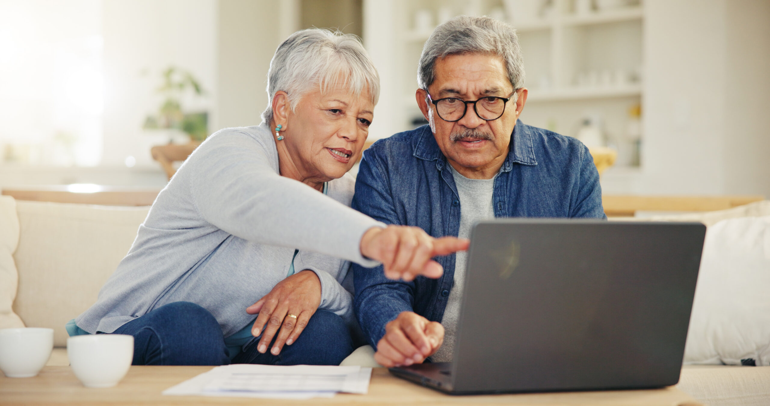 Older couple sitting on a sofa, looking at a laptop together as they point at the screen.