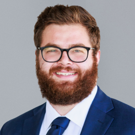 Professional headshot of a bearded man wearing glasses in a navy suit and tie, smiling against a gray background.
