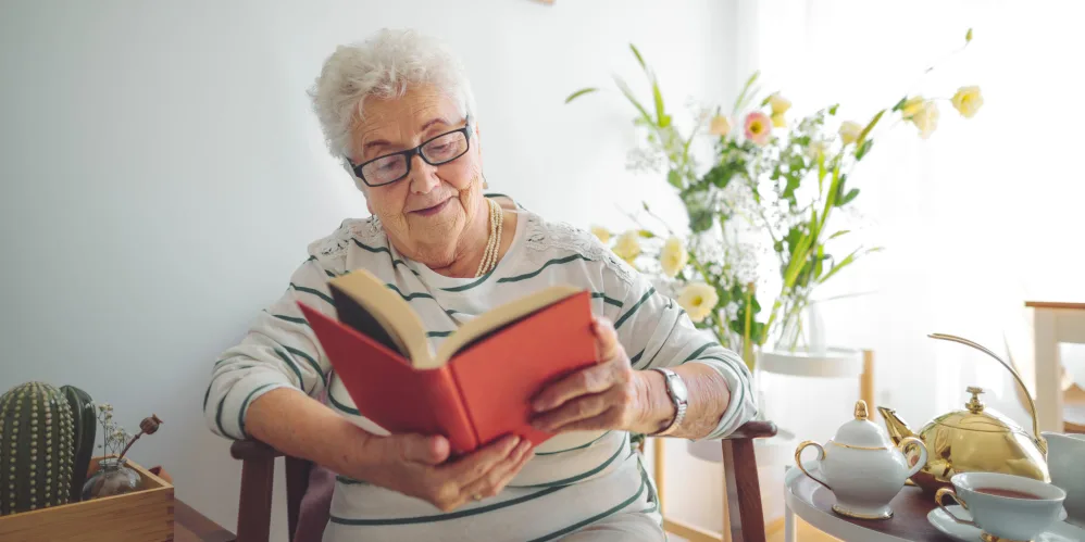 Serene elderly woman enjoying reading a book at Vista Prairie at Copperleaf senior living facility in Willmar, MN, with bright natural light and fresh flowers showcasing comfort and quality care.