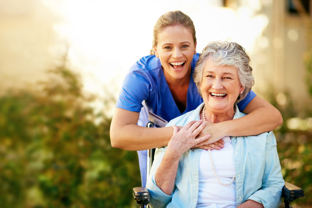 Elderly woman in wheelchair with smiling caregiver outdoors in sunlight, promoting senior living and assisted care services at Vista Prairie at Copperleaf.