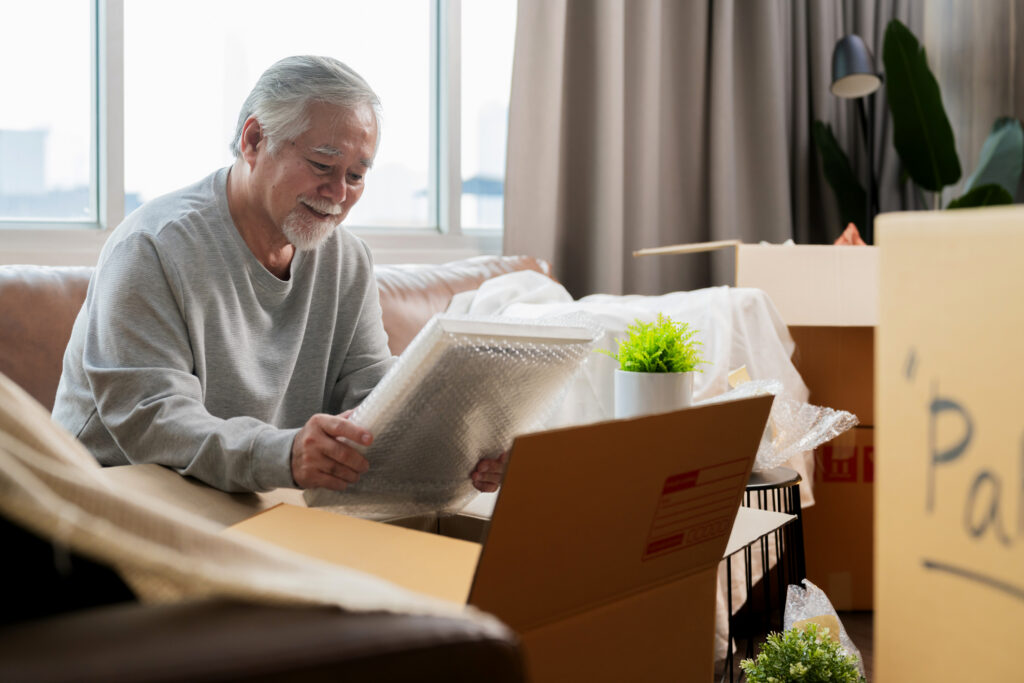 senior man surrounded by moving boxes, looking at picture frame in bubble wrap