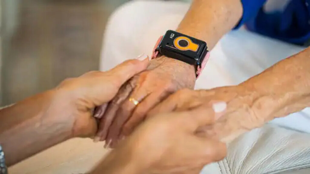 Caregiver supports an elderly person’s wrist while wearing a black smartwatch with an orange dial. Two hands grip an elderly forearm; a black smartwatch with an orange circle is on the wrist. Close-up of hands holding an elderly wrist with a bright orange-ring smartwatch. Caregiver assisting an elderly person; smartwatch visible on the forearm.