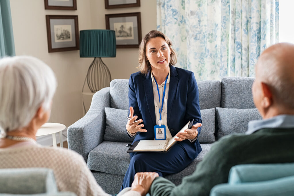 Professional woman in a blue suit speaking to a group in a living room, holding a notebook and pen with a notepad on her lap.