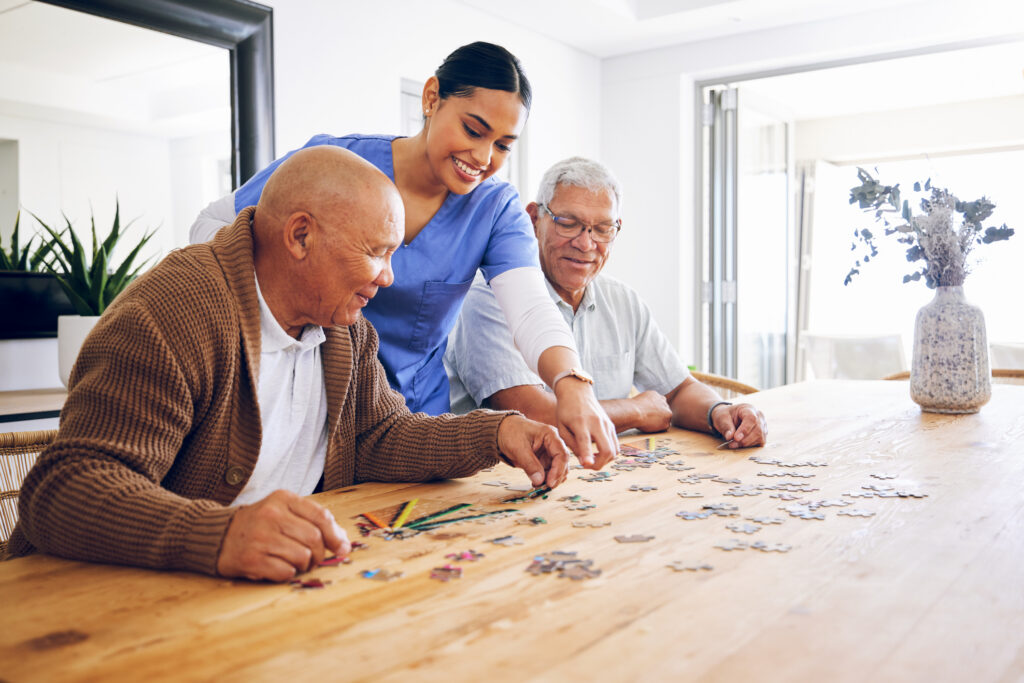 Caregiver in blue scrubs helping two elderly men assemble a puzzle at a wooden table in a bright room, smiling and collaborating.