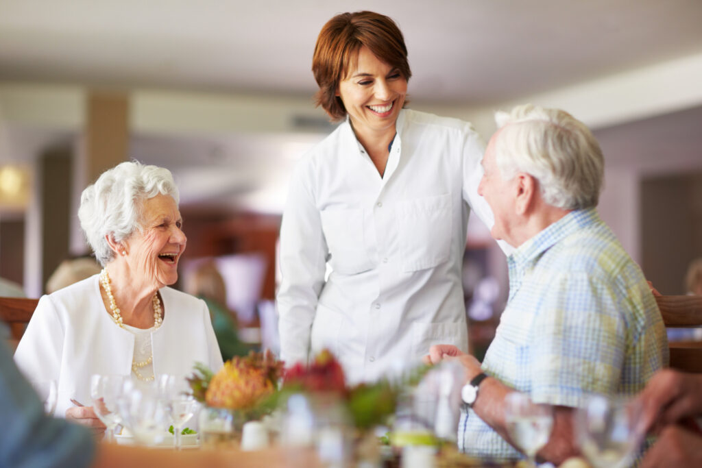 Caregiver smiling at two elderly diners during a meal in a bright dining area.