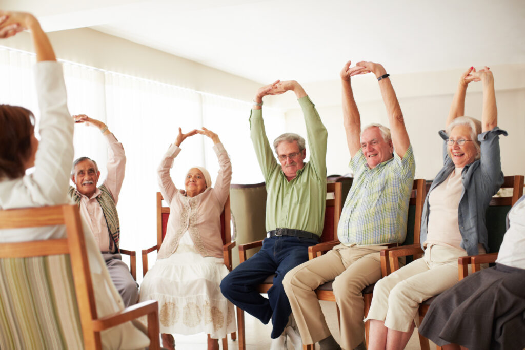 Group of elderly adults seated in chairs, raising their arms in a stretching exercise in a bright room.